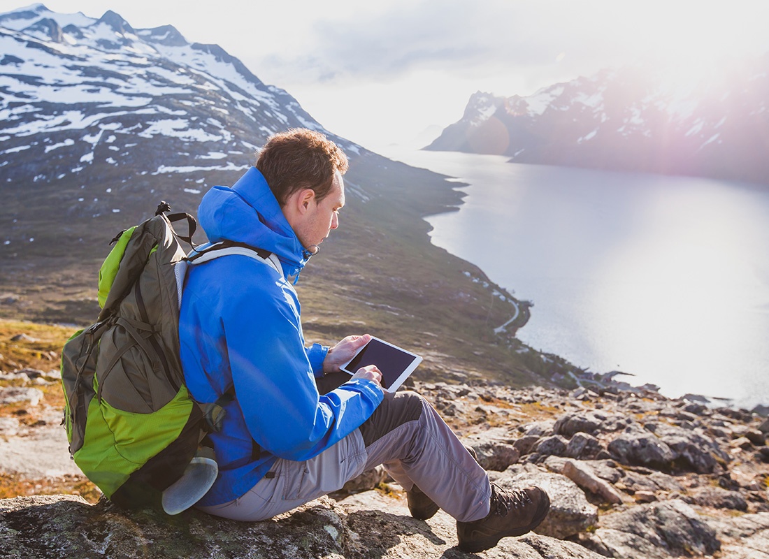 Service Center - Man Sits Down on a Rocky Mountain by a Lake and Uses His Tablet